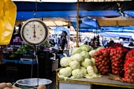 A bustling outdoor market scene with a hanging scale in the foreground, surrounded by fresh produce like cabbages and potatoes. Stalls are covered with blue and yellow tarps, and people are walking and interacting in the background.