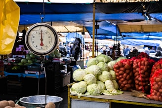 A bustling outdoor market scene with a hanging scale in the foreground, surrounded by fresh produce like cabbages and potatoes. Stalls are covered with blue and yellow tarps, and people are walking and interacting in the background.