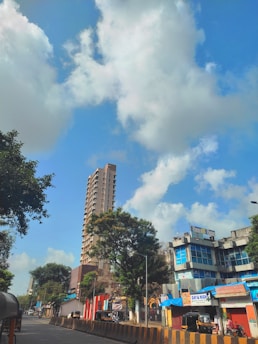 A busy urban street scene with a tall residential building and a smaller commercial building with blue signage. The sky is bright and filled with large white clouds. Several trees line the street, and a few vehicles, including auto rickshaws, are parked or moving along the road.