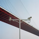 Two surveillance cameras mounted on a white pole, set against the backdrop of the underside of a large red suspension bridge with cables stretching upwards. The sky is clear and blue, providing a contrast to the red and white structures.