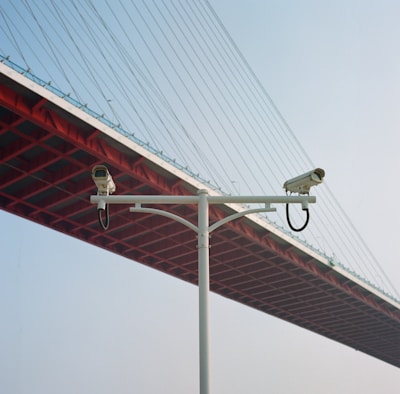 Two surveillance cameras mounted on a white pole, set against the backdrop of the underside of a large red suspension bridge with cables stretching upwards. The sky is clear and blue, providing a contrast to the red and white structures.