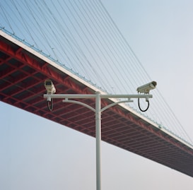 Two surveillance cameras mounted on a white pole, set against the backdrop of the underside of a large red suspension bridge with cables stretching upwards. The sky is clear and blue, providing a contrast to the red and white structures.