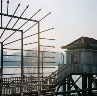 An ornate metal fence with spear-like points outlines the foreground, partially obscuring a view of a body of water with shimmering reflections. A building with a tiled roof and balcony, connected by a staircase, stands near the fence. Indistinct skyscrapers are visible in the distant background, giving a sense of a sunset or sunrise setting.