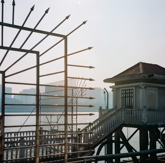 An ornate metal fence with spear-like points outlines the foreground, partially obscuring a view of a body of water with shimmering reflections. A building with a tiled roof and balcony, connected by a staircase, stands near the fence. Indistinct skyscrapers are visible in the distant background, giving a sense of a sunset or sunrise setting.