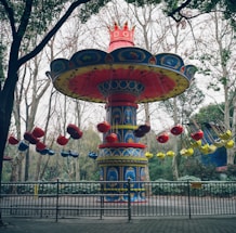 A brightly colored, elaborate amusement park ride with ornate decorations. The ride has a large central column with a red crown-like top, and circular seating arrangements suspended by chains. The seats are predominantly red, blue, and yellow. The ride is surrounded by trees and fenced off by a metallic barricade.