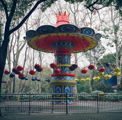 A brightly colored, elaborate amusement park ride with ornate decorations. The ride has a large central column with a red crown-like top, and circular seating arrangements suspended by chains. The seats are predominantly red, blue, and yellow. The ride is surrounded by trees and fenced off by a metallic barricade.