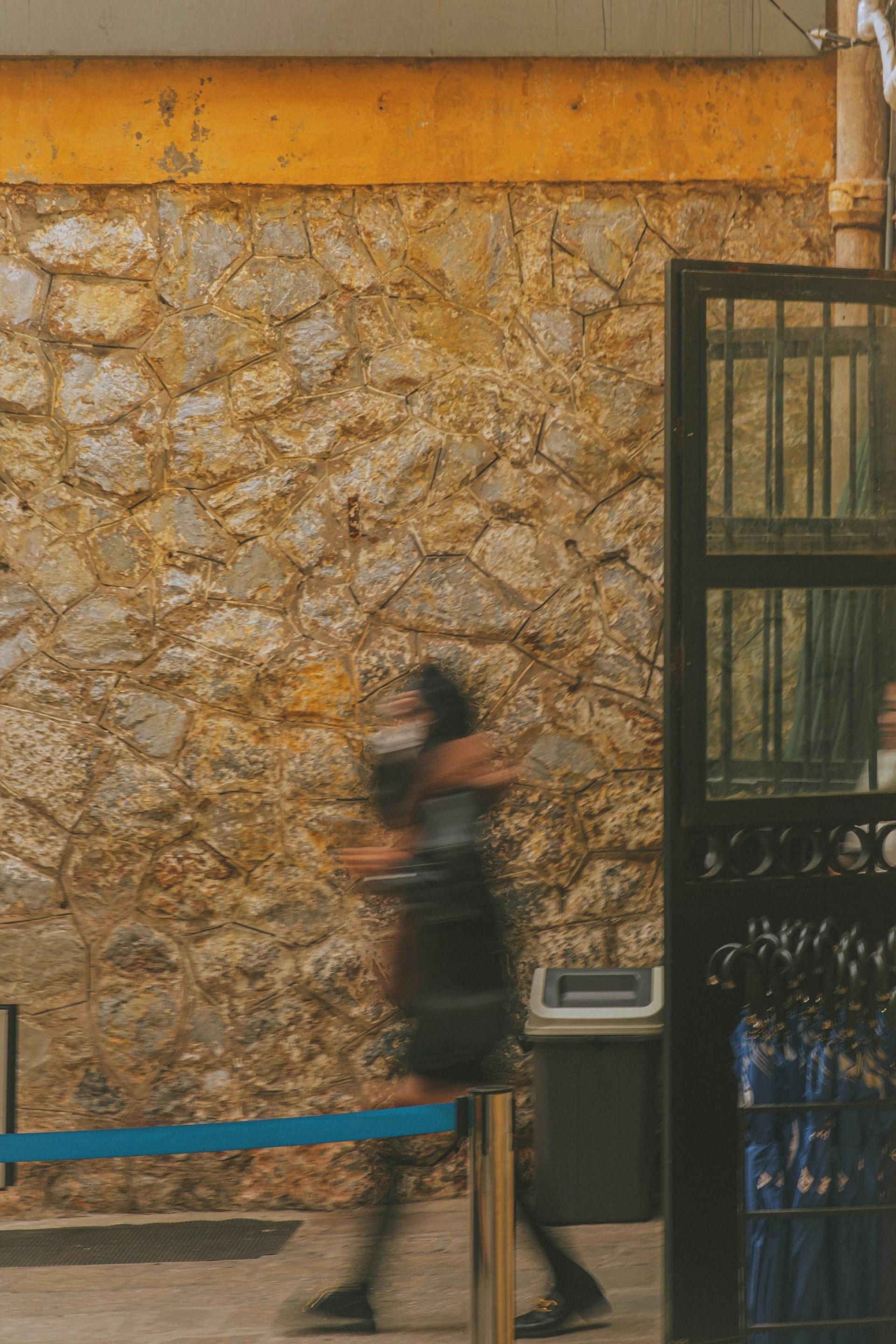 A person wearing a mask is walking quickly past a textured stone wall. The motion creates a slight blur effect. A trash can is situated near the wall, close to an open black metal gate. The scene gives an impression of haste, with the contrasting colors of the textured yellow and brown wall.
