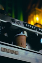 A close-up of a coffee machine with a takeaway cup labeled 'Juggernaut Cold Coffee' resting on the tray. The machine has a silver and black finish with buttons and a pressure gauge. Warm yellow lighting in the background adds a cozy ambiance.