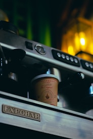 A close-up of a coffee machine with a takeaway cup labeled 'Juggernaut Cold Coffee' resting on the tray. The machine has a silver and black finish with buttons and a pressure gauge. Warm yellow lighting in the background adds a cozy ambiance.