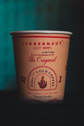 A paper cup with branding and text featuring the word 'Juggernaut' prominently at the top. The design includes the phrase 'ESTP 2017½' and references to 'The Original' and 'COLD COFFEE'. The cup has decorative elements and smaller text in various languages and fonts, giving it a stylish and artisanal feel.