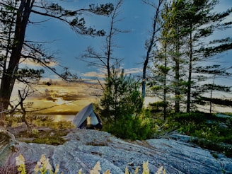 A serene camping scene with a tent set up on rocky ground amidst tall pine trees. The sun is setting over a lake in the background, casting a warm glow across the landscape. The sky is a mix of blue and orange hues.