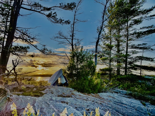 A serene camping scene with a tent set up on rocky ground amidst tall pine trees. The sun is setting over a lake in the background, casting a warm glow across the landscape. The sky is a mix of blue and orange hues.