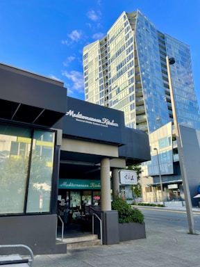 The image features an urban street scene with a modern high-rise building in the background. In the foreground, there is a restaurant with a sign displaying 'Mediterranean Kitchen' and 'Gourmet Middle Eastern Cuisine'. The structure is contemporary with large glass windows, and there's another sign for 'Santouka'. The sky is clear with a few clouds, and the scene is well-lit, suggesting it's daytime.