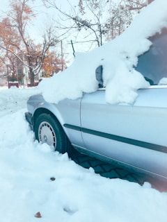 A car is parked along a street, covered with a thick layer of snow. Snow blankets the windshield and hood, and the vehicle's roof is similarly topped with snow. Surrounding trees are bare, suggesting a winter setting. Nearby, other parts of the street are also covered in snow.
