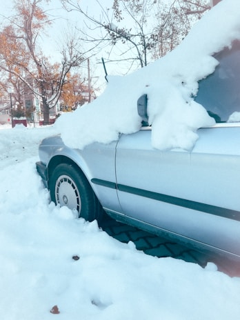 A car is parked along a street, covered with a thick layer of snow. Snow blankets the windshield and hood, and the vehicle's roof is similarly topped with snow. Surrounding trees are bare, suggesting a winter setting. Nearby, other parts of the street are also covered in snow.