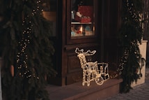 A festive storefront is adorned with pine garlands and small golden lights. An illuminated reindeer decoration is placed at the entrance, with a window display featuring wrapped gifts and ornaments.