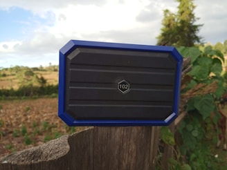 A blue-framed hexagonal speaker with a number 102 on the front is positioned on a wooden post in an outdoor rural setting. The background features a field with sparse vegetation and trees under a partly cloudy sky.