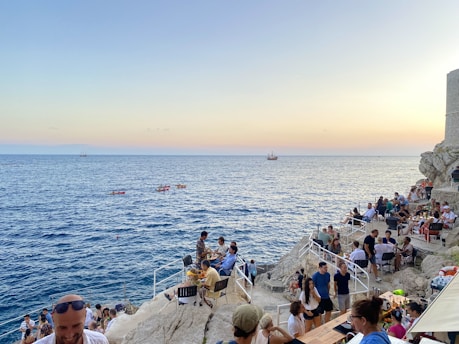 People are enjoying a seaside bar or cafe located on rocky cliffs overlooking the ocean. The sky displays pastel colors as the sun sets, and several boats or kayaks are visible on the water. The scene is vibrant with numerous individuals sitting, walking, and chatting, creating a lively atmosphere.