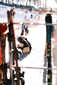 A ski resort scene with several people wearing winter gear. A person is sitting on the ground, possibly adjusting ski equipment. Ski poles or skis are visible in the foreground, slightly out of focus. The background shows snow, safety netting, and trees, with other visitors in the distance.