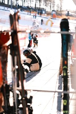 A ski resort scene with several people wearing winter gear. A person is sitting on the ground, possibly adjusting ski equipment. Ski poles or skis are visible in the foreground, slightly out of focus. The background shows snow, safety netting, and trees, with other visitors in the distance.