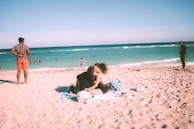 A sunny beach scene with people enjoying the sand and sea. A couple sits on a blanket in the foreground, embracing and surrounded by bags and beach items. Behind them, several people stand or walk along the shoreline, with the turquoise ocean and clear sky providing a serene backdrop.