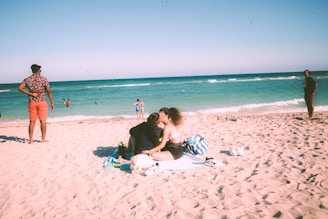 A sunny beach scene with people enjoying the sand and sea. A couple sits on a blanket in the foreground, embracing and surrounded by bags and beach items. Behind them, several people stand or walk along the shoreline, with the turquoise ocean and clear sky providing a serene backdrop.