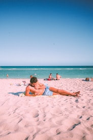 A relaxed beach scene with several people enjoying the sunny day. In the foreground, a man lies on a towel on the sandy beach, looking at his phone. Other people in the distance are sitting and chatting near the water. The ocean is calm, and the sky is clear, creating a peaceful atmosphere.