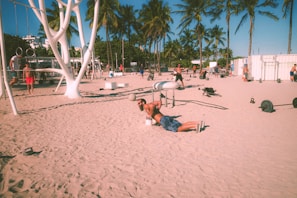 People are exercising on a sandy beach with a variety of gym equipment. Palm trees line the background, and the atmosphere suggests a warm, sunny day. Individuals are engaged in different workouts, including weightlifting and calisthenics.