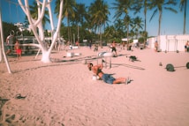 People are exercising on a sandy beach with a variety of gym equipment. Palm trees line the background, and the atmosphere suggests a warm, sunny day. Individuals are engaged in different workouts, including weightlifting and calisthenics.