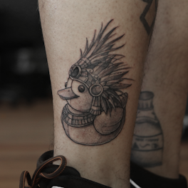 A tattoo of a rubber duck with an intricate headdress resembling tribal or Aztec designs, featuring feathers and circular details. The tattoo is located on a person's leg.