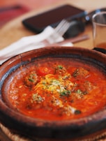 A dish featuring meatballs in a rich, red sauce garnished with herbs. The food is served in a rustic, round clay serving dish placed on a wooden surface, with a fork and knife in the background.