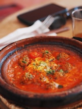 A dish featuring meatballs in a rich, red sauce garnished with herbs. The food is served in a rustic, round clay serving dish placed on a wooden surface, with a fork and knife in the background.