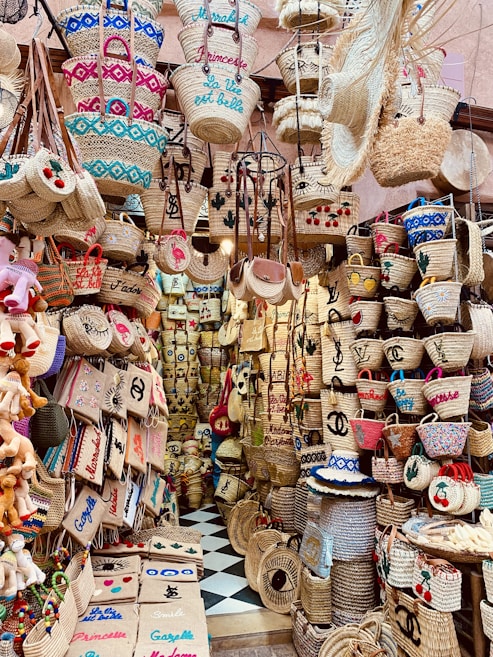 A vibrant market stall displays a wide array of woven straw baskets of various shapes and sizes. Many baskets have colorful embroidery and designs featuring words, symbols, and patterns. The setting is densely packed with items stacked on shelves and hanging on hooks, creating a visually striking and busy atmosphere.