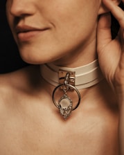 A close-up of a person's neck with a beige choker featuring a large metallic ring and a decorative pendant.
