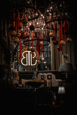A dimly lit interior space featuring a decorative chandelier adorned with hanging ornaments and ribbons. Below the chandelier, there is a table with numbered boxes, a bowl of wrapped items, and decorative items. A round sign with stylized letters is displayed prominently.