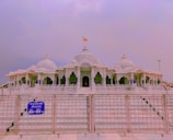 An intricately designed temple with a series of domes and arches made from white marble. The temple features detailed carvings and a prominent flag atop the central dome. A metal gate and sign with blue background and white text are positioned in the foreground.
