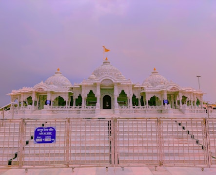An intricately designed temple with a series of domes and arches made from white marble. The temple features detailed carvings and a prominent flag atop the central dome. A metal gate and sign with blue background and white text are positioned in the foreground.