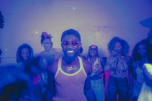 A group of people are gathered in a dimly lit room with purple and blue lighting. One person at the center is smiling brightly and wearing a mesh tank top and sunglasses. Others around are dressed in casual attire, some holding drinks, and appear to be enjoying a social gathering.
