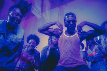 A group of people appear to be enjoying themselves in a lively, colorful environment. A man in the center strikes a pose with his arms flexed, wearing sunglasses and a mesh tank top, while others around him are smiling and cheering. The lighting casts a purple and blue hue over the scene, adding to the energetic and dynamic atmosphere.