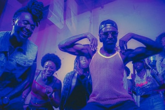 A group of people appear to be enjoying themselves in a lively, colorful environment. A man in the center strikes a pose with his arms flexed, wearing sunglasses and a mesh tank top, while others around him are smiling and cheering. The lighting casts a purple and blue hue over the scene, adding to the energetic and dynamic atmosphere.