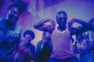 A group of people appear to be enjoying themselves in a lively, colorful environment. A man in the center strikes a pose with his arms flexed, wearing sunglasses and a mesh tank top, while others around him are smiling and cheering. The lighting casts a purple and blue hue over the scene, adding to the energetic and dynamic atmosphere.