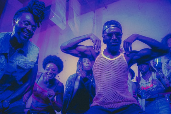 A group of people appear to be enjoying themselves in a lively, colorful environment. A man in the center strikes a pose with his arms flexed, wearing sunglasses and a mesh tank top, while others around him are smiling and cheering. The lighting casts a purple and blue hue over the scene, adding to the energetic and dynamic atmosphere.