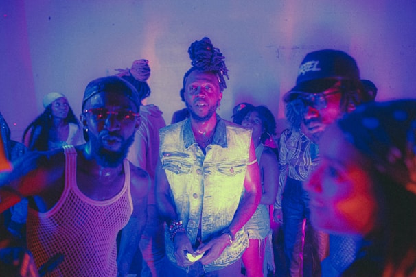 A group of people is gathered in a dimly lit setting with a vibrant mix of purple and blue lighting. The central figure, wearing a sleeveless denim jacket and adorned with a stylish dreadlock bun, looks directly at the camera. Other individuals are casually dressed, some wearing hats, and are engaged in various activities, creating an energetic and lively atmosphere.