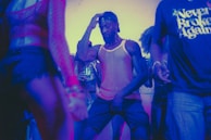 A group of people dancing in a dimly lit environment with colorful lighting. One person is at the center of the image, wearing a mesh tank top and a backward cap, deeply immersed in dance. The surrounding individuals are also engaged in dancing or standing nearby, some with partially visible faces and clothing.