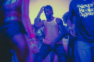 A group of people dancing in a dimly lit environment with colorful lighting. One person is at the center of the image, wearing a mesh tank top and a backward cap, deeply immersed in dance. The surrounding individuals are also engaged in dancing or standing nearby, some with partially visible faces and clothing.