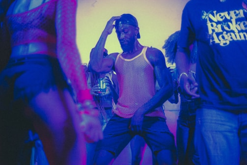A group of people dancing in a dimly lit environment with colorful lighting. One person is at the center of the image, wearing a mesh tank top and a backward cap, deeply immersed in dance. The surrounding individuals are also engaged in dancing or standing nearby, some with partially visible faces and clothing.