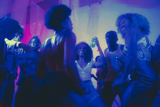 A lively group of people dancing at a party with vibrant lighting and a festive atmosphere. Most individuals are holding drinks, and the setting is filled with colorful lights, giving a neon-like effect.