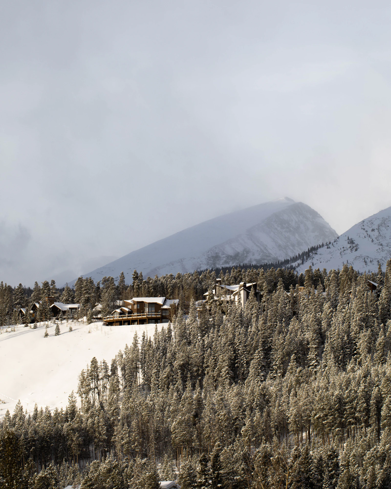 A snow-covered mountainside with a ski lodge in the distance under low cloud