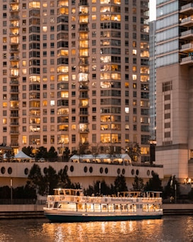 A sightseeing boat illuminated with warm lights floats on a calm waterway in front of tall, modern high-rise buildings. The windows of the buildings are lit, suggesting evening or night time, with reflections visible on the water. Trees and architectural elements are seen along the waterfront.
