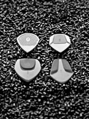 Four silver guitar picks are placed on a bed of small black cylindrical pellets. The picks are arranged in a neat pattern, showcasing various textures and designs on each.